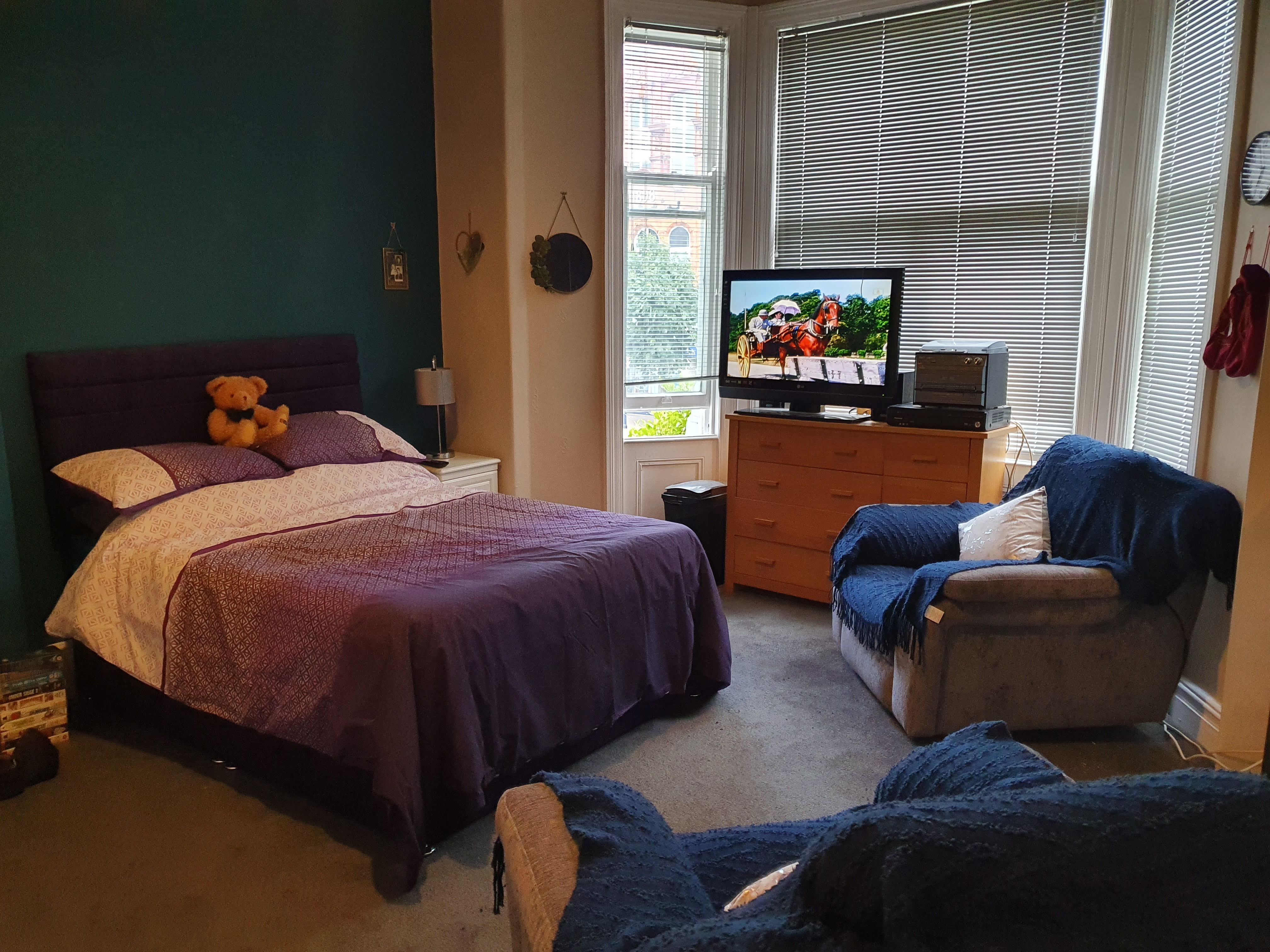 The image shows: A bedroom with a purple bed, a TV on a wooden cabinet in front of a large window, and a armchair with a m...